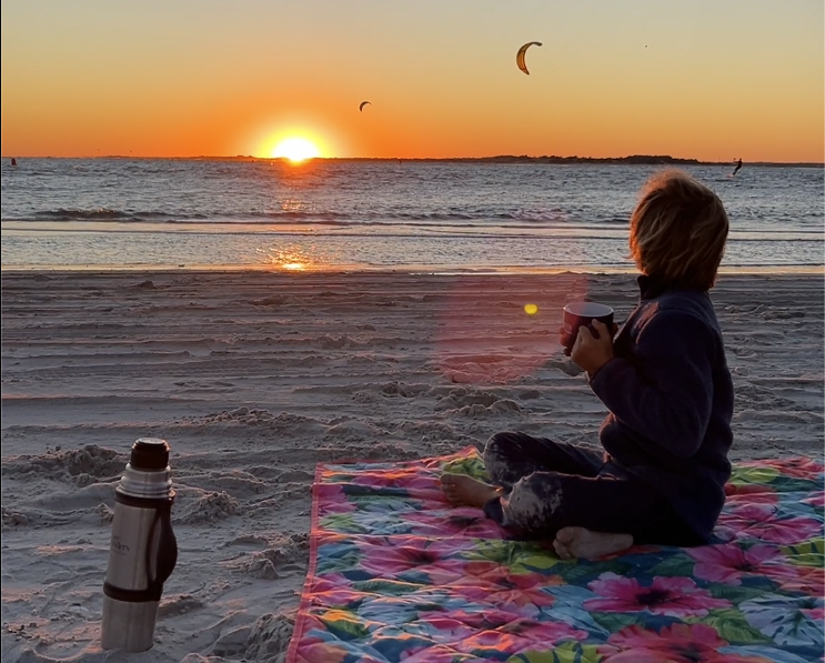 Child drinking hot chocolate at sunset with kitesurfers in the background